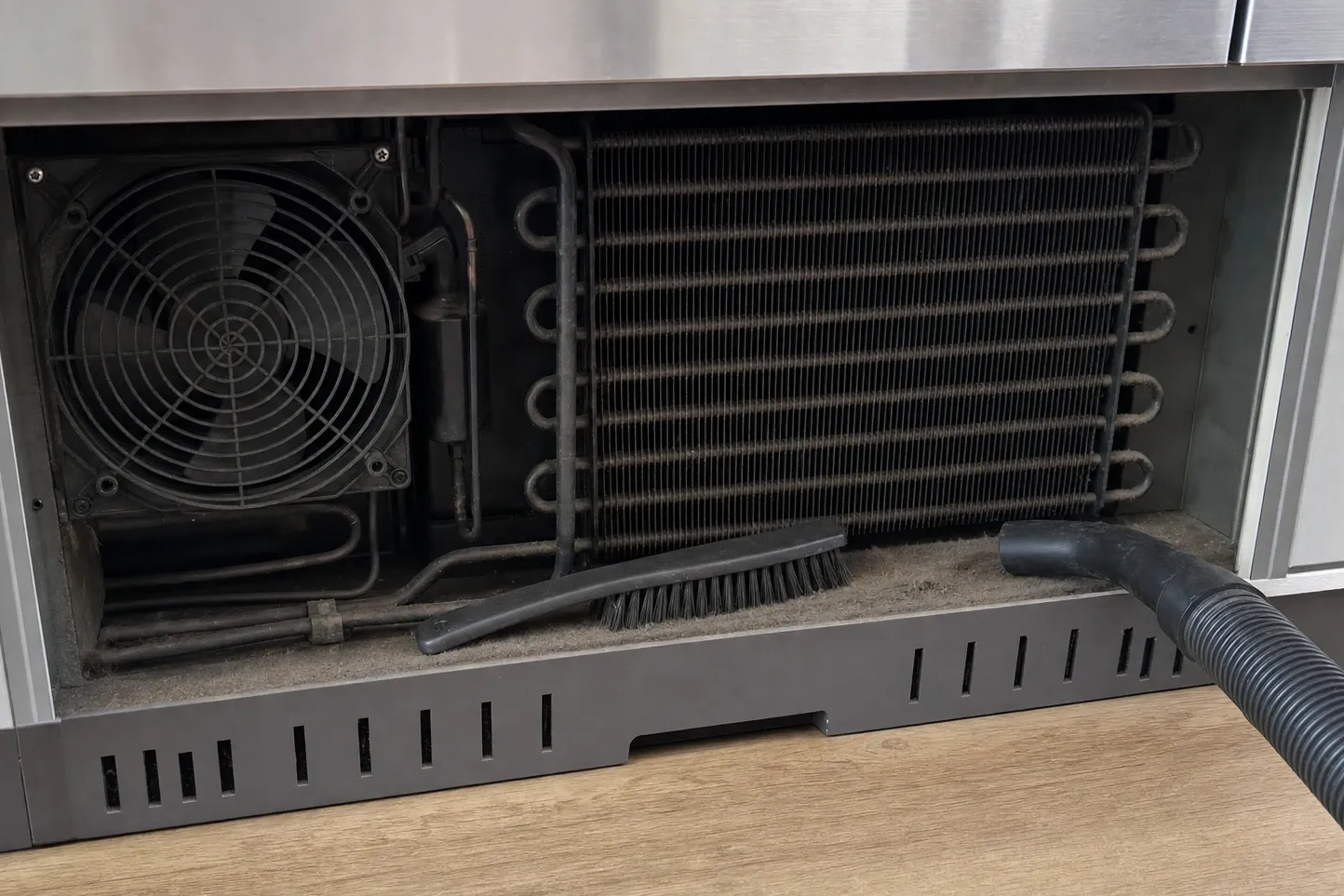 Lower grille removed from a stainless built-in refrigerator, exposing a condenser coil matted with grey lint, with a brush and vacuum nozzle on the floor